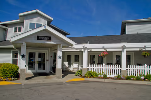 Exterior view of a senior living facility building with a covered entrance, white picket fence, hanging flower baskets, and clear blue sky.