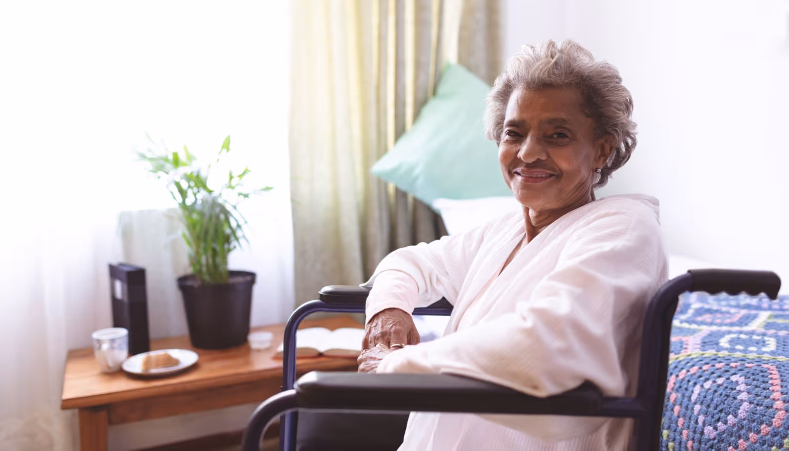 An elderly woman sitting in a wheelchair in a cozy room with a bed covered by a colorful crocheted blanket. There is a wooden side table next to her with a potted plant, a plate with a pastry, a cup, and an open book. Light filters through a window with curtains in the background.