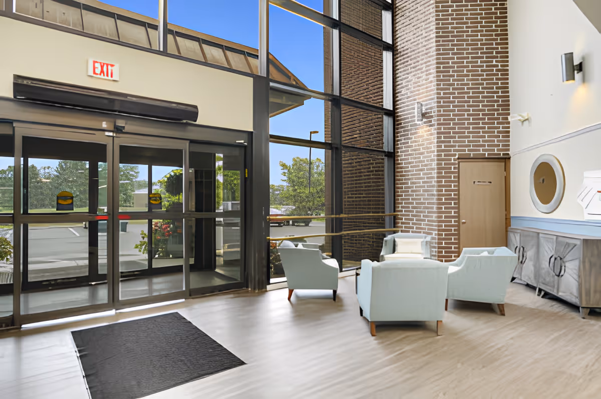 Sunny entrance lobby with automatic glass doors, tall floor-to-ceiling windows, a brick accent wall, and a small seating area of light blue chairs.