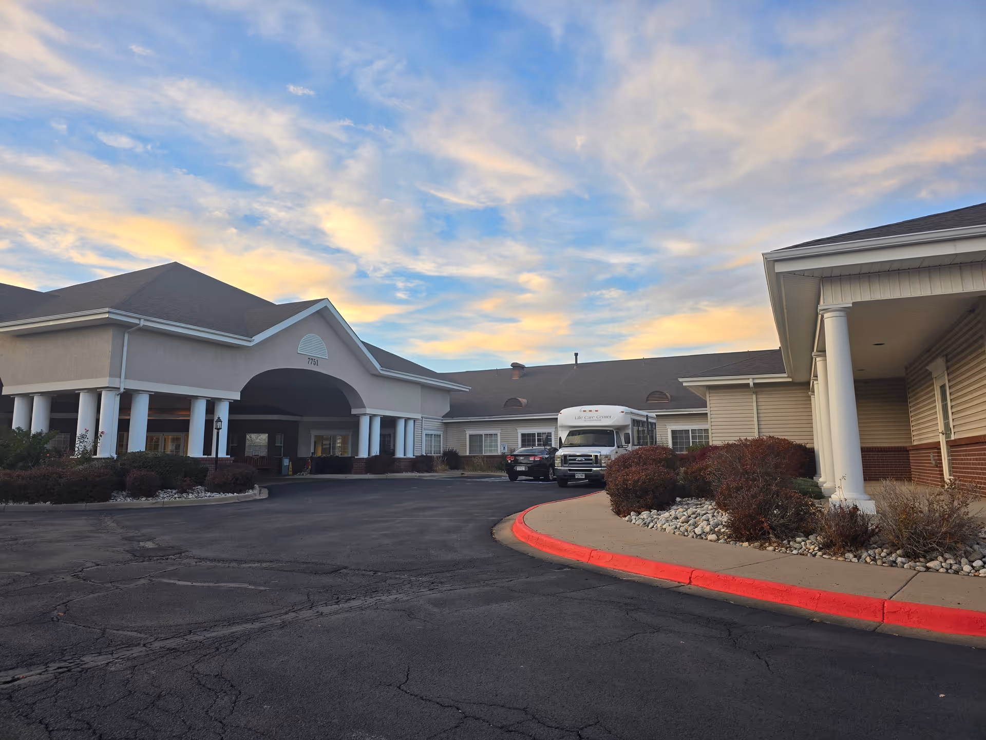 Exterior front entrance of a retirement community with a covered porte-cochère, landscaped beds, driveway, and a parked shuttle van under a colorful sky.