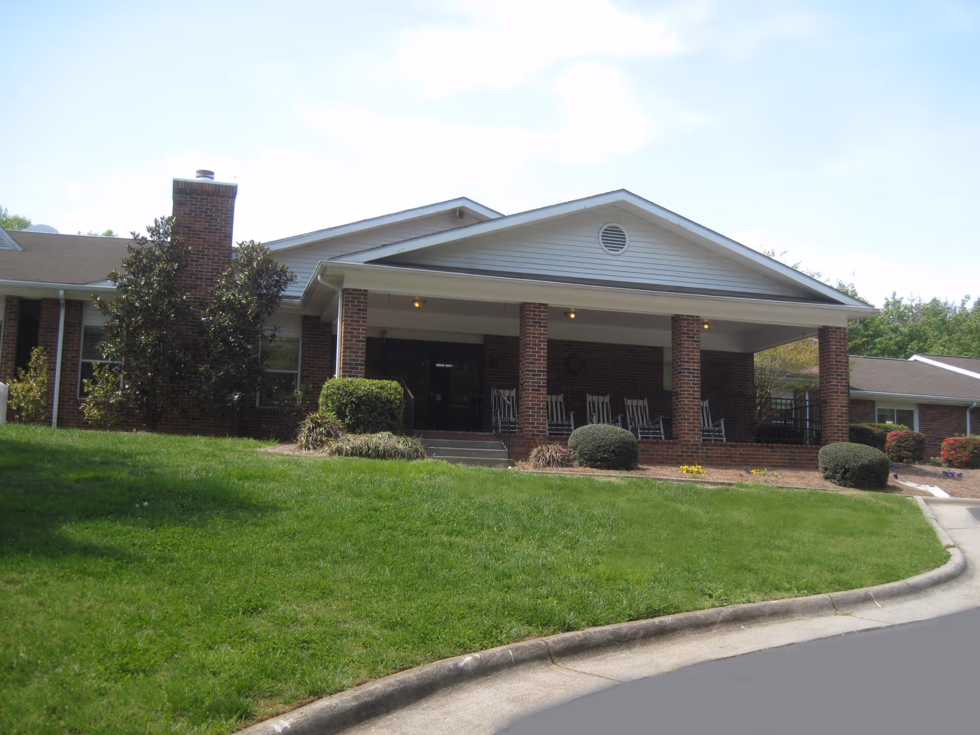 Front entrance of a brick senior living facility with a covered porch, rocking chairs, and a grassy lawn.