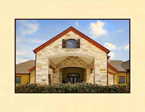 Stone facade entrance of a senior living facility with shrubs and blue sky.