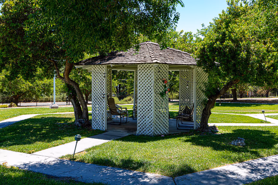 A white wooden gazebo with lattice walls and a shingled roof sits in a green grassy area surrounded by trees and paved walkways under a clear blue sky.