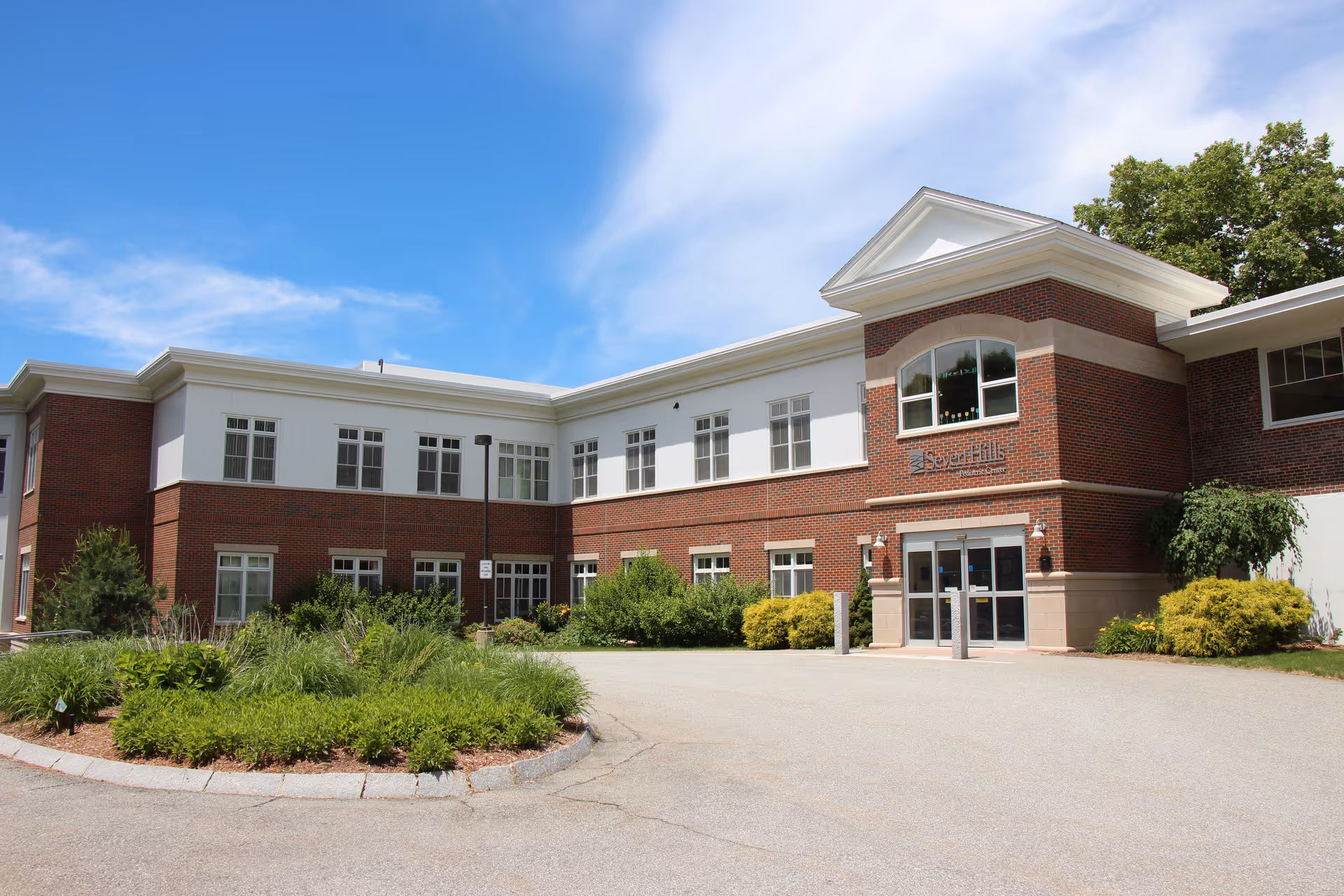 Exterior view of a two-story brick and white panel building under a blue sky with some clouds. The building has multiple windows and a main entrance with glass doors. There is a landscaped area with green shrubs and plants in front of the building.