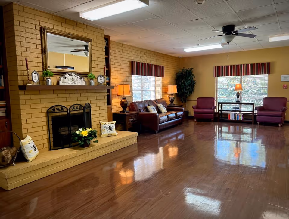 A warm communal living room with a brick fireplace, leather sofa, upholstered armchairs, lamps, and a shiny hardwood floor.