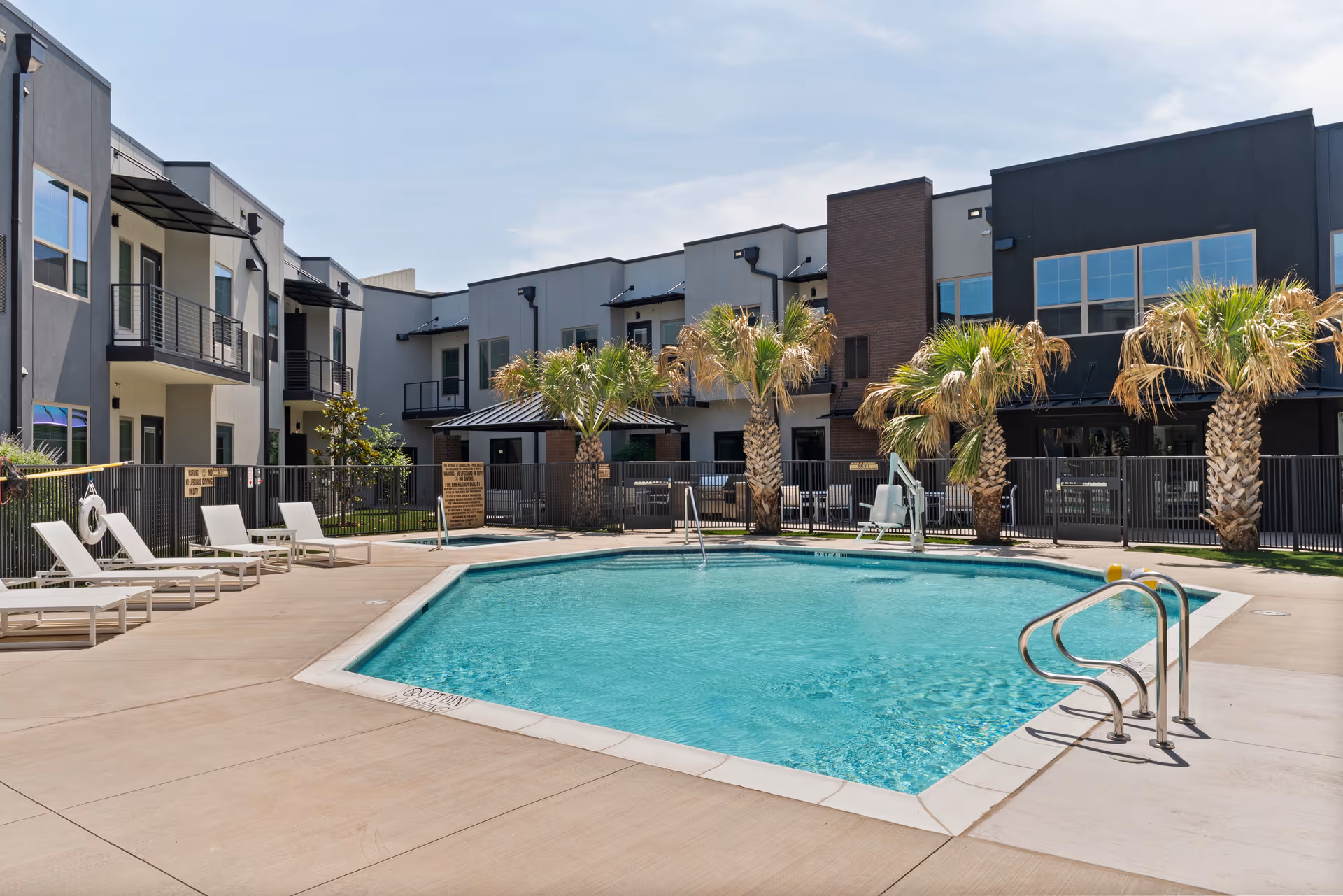 Outdoor swimming pool surrounded by a concrete deck with several white lounge chairs. The pool area is fenced and has palm trees planted along the fence. Modern two-story buildings with balconies and large windows are visible in the background under a clear sky.