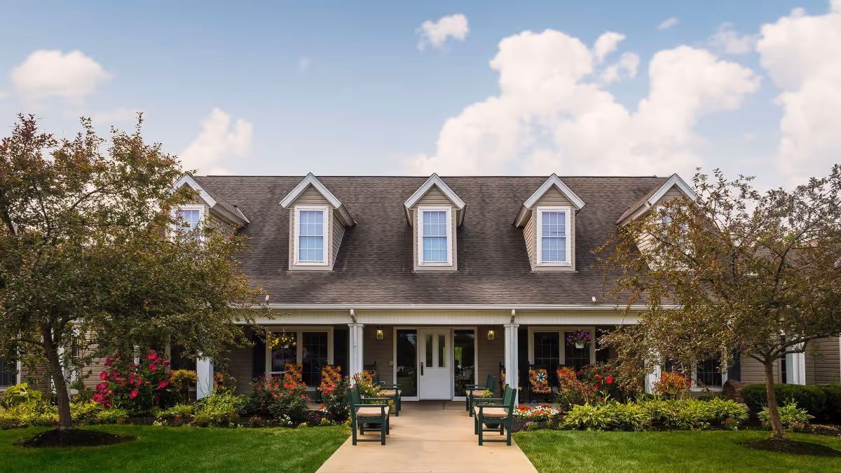 Front exterior view of a residential building with a porch, green lawn, trees, and flower beds under a partly cloudy sky.