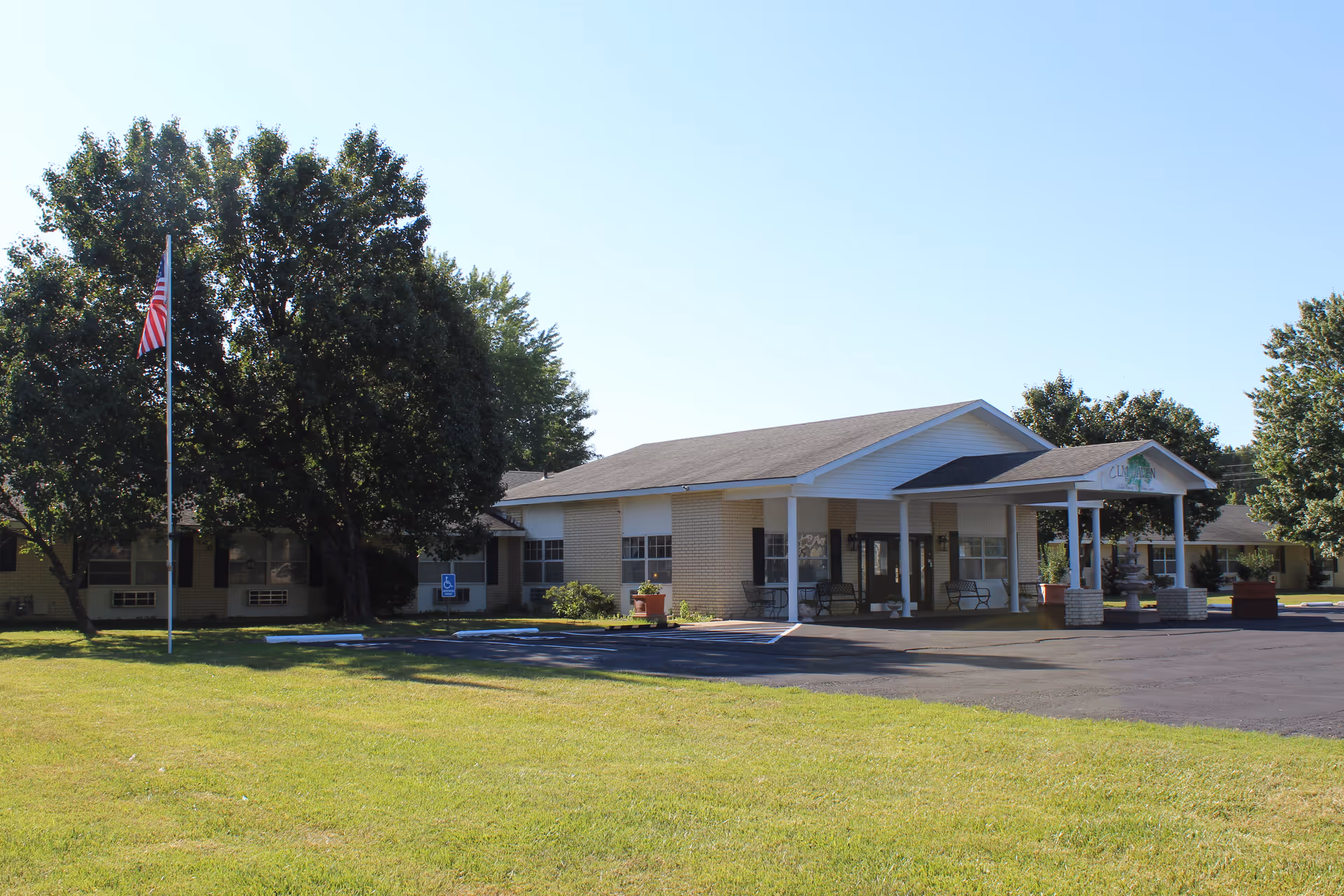 Single-story brick senior living facility with a covered entrance, flagpole, and lawn with trees.