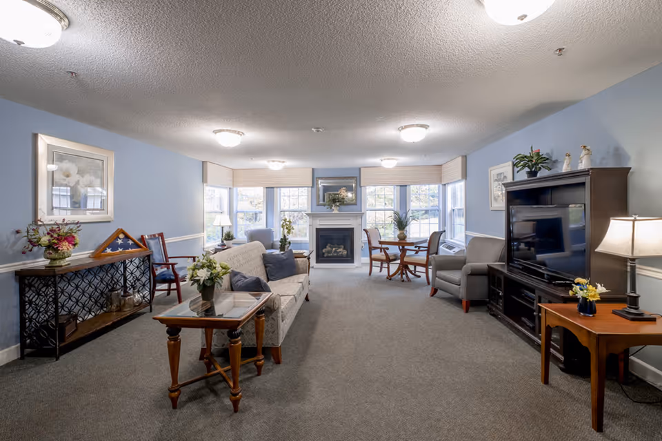 A cozy living room with light blue walls and carpeted floor. The room features a white sofa with blue cushions, a wooden coffee table with a flower vase, a fireplace centered between three large windows with beige blinds, a round wooden table with four chairs near the windows, a gray armchair, and a dark wooden TV stand with a flat-screen television. Decorative items include framed artwork, plants, and lamps providing warm lighting.