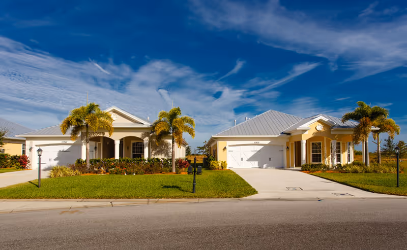 Two single-story pastel-colored houses with palm trees, garages and driveways under a bright blue sky.