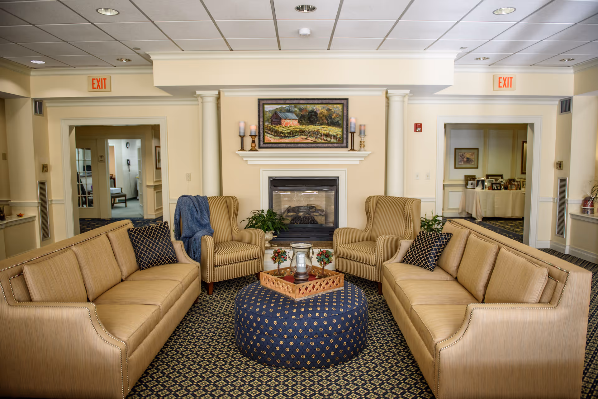 Comfortable senior living room lounge with two sofas and armchairs arranged around a central patterned ottoman in front of a fireplace.