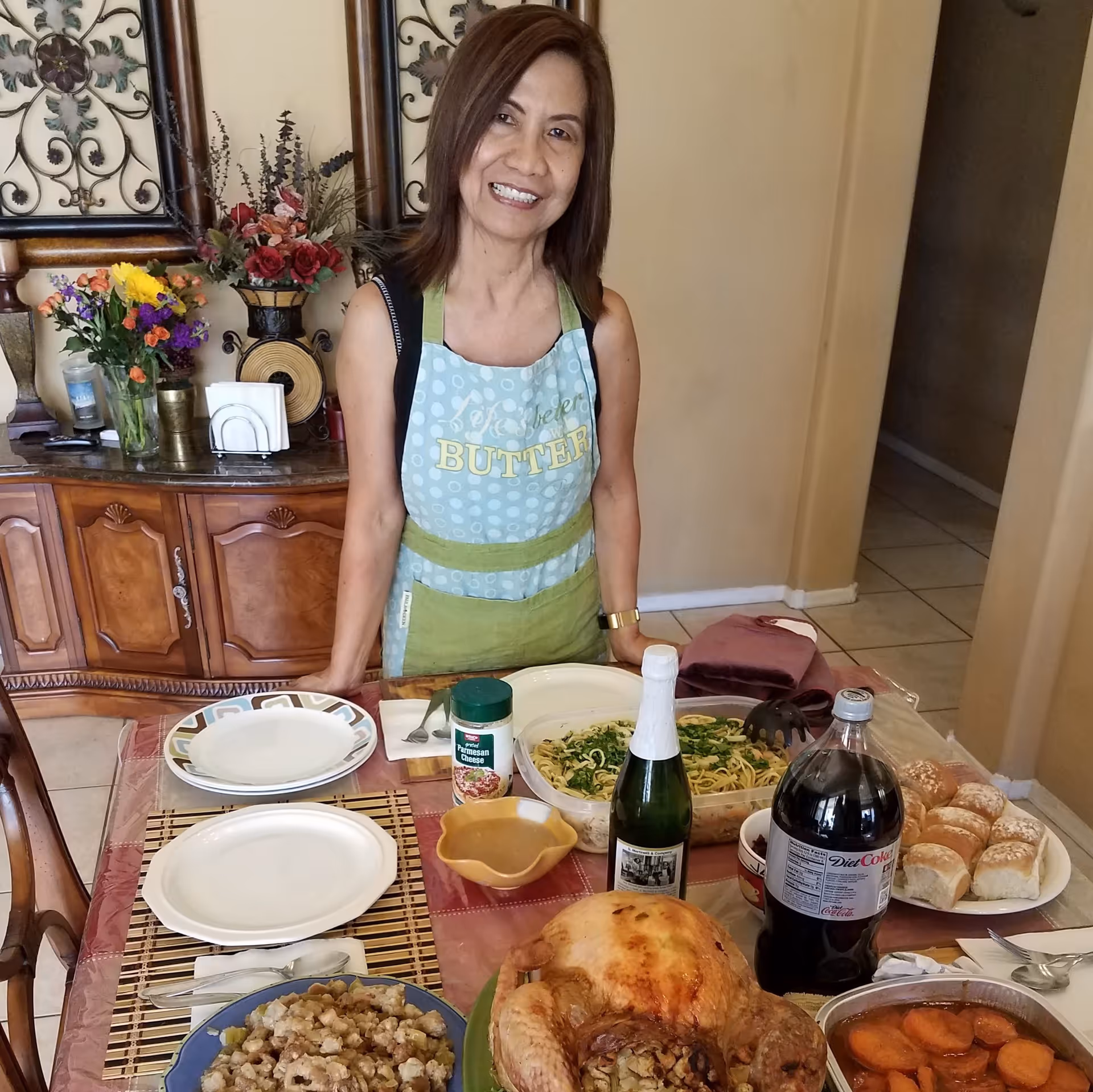 A woman wearing an apron stands behind a dining table set with various dishes including a roasted turkey, stuffing, pasta, dinner rolls, a bottle of Diet Coke, and a bottle of sparkling beverage. The background shows a wooden sideboard with flower arrangements and decorative wall art.