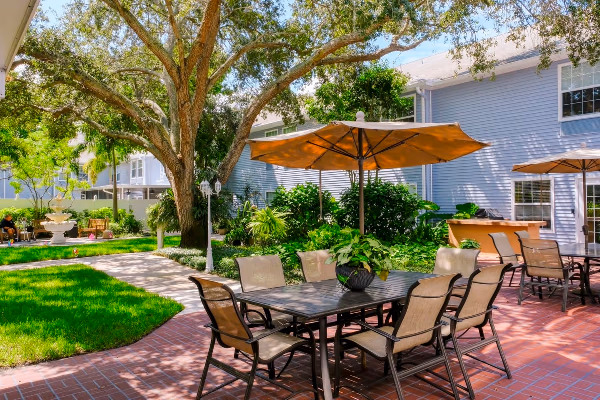 Outdoor courtyard patio with tables, umbrellas and chairs under a large tree beside a residential building and fountain.