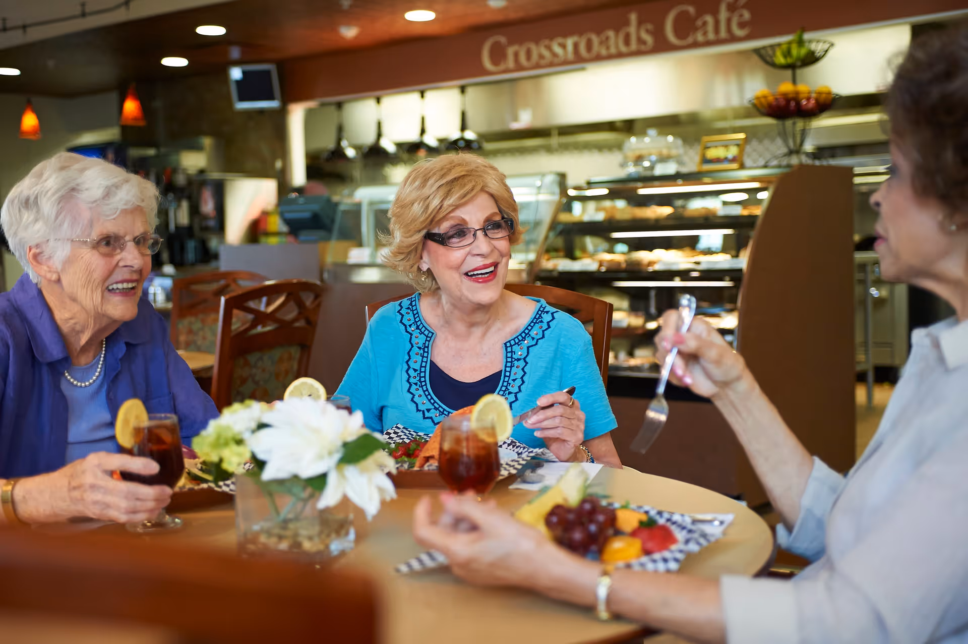Three elderly women sitting around a table in a café, enjoying a meal and drinks. The table has plates of food including fruit and iced tea with lemon slices. In the background, there is a display case with baked goods and a sign that reads 'Crossroads Café'.