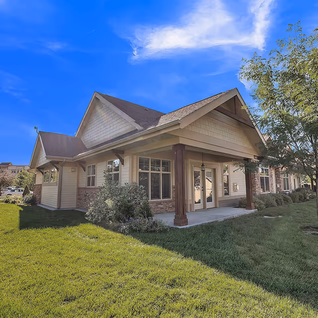 Exterior view of a single-story building with a covered porch supported by wooden columns, surrounded by green grass and trees under a bright blue sky.