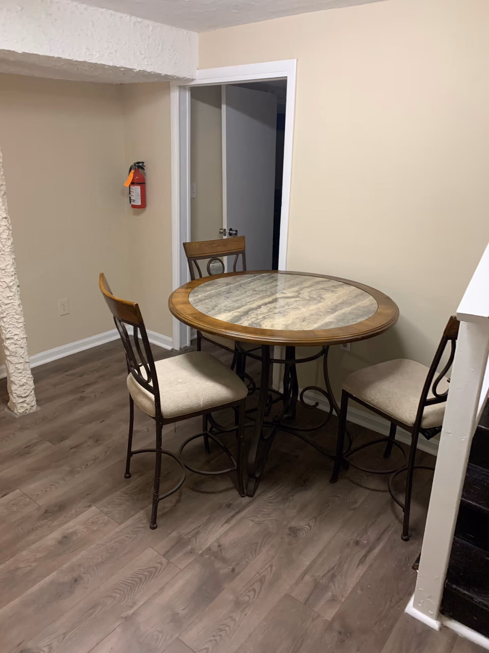 A small dining area with a round table featuring a marble-like surface and wooden trim, surrounded by three cushioned chairs with metal frames. The room has light beige walls, wood-patterned flooring, a fire extinguisher mounted on the wall, and an open doorway leading to another room.