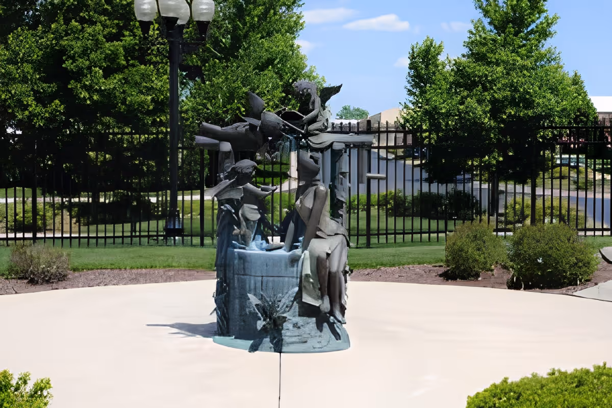 Outdoor sculpture of a woman and child sitting by a well, surrounded by greenery, trees, and a black metal fence under a blue sky with some clouds.