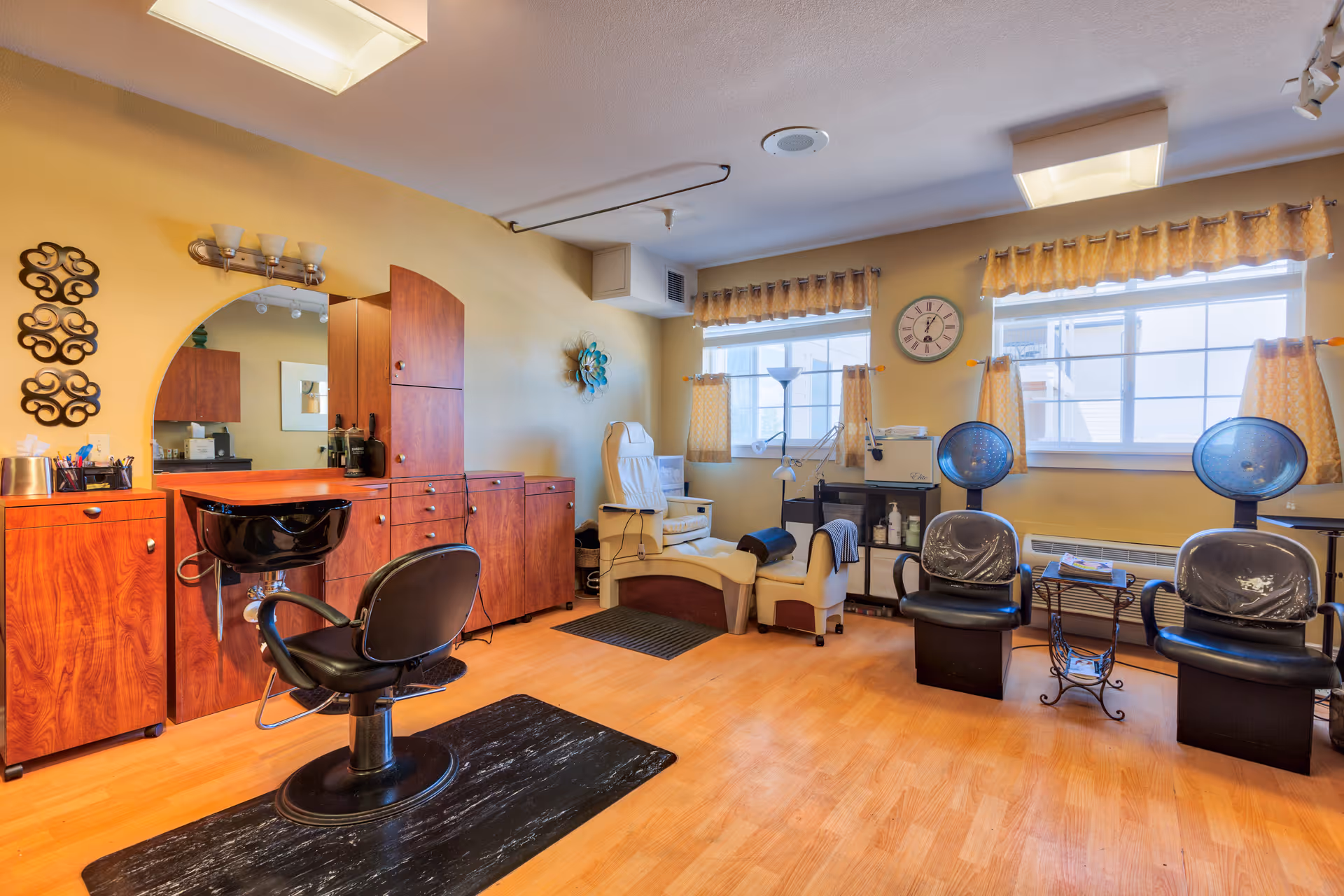 Interior view of a senior living facility's salon area with wooden cabinets, a black salon chair in front of a wash basin, two black hair drying chairs, a white reclining chair, and windows with yellow curtains letting in natural light.