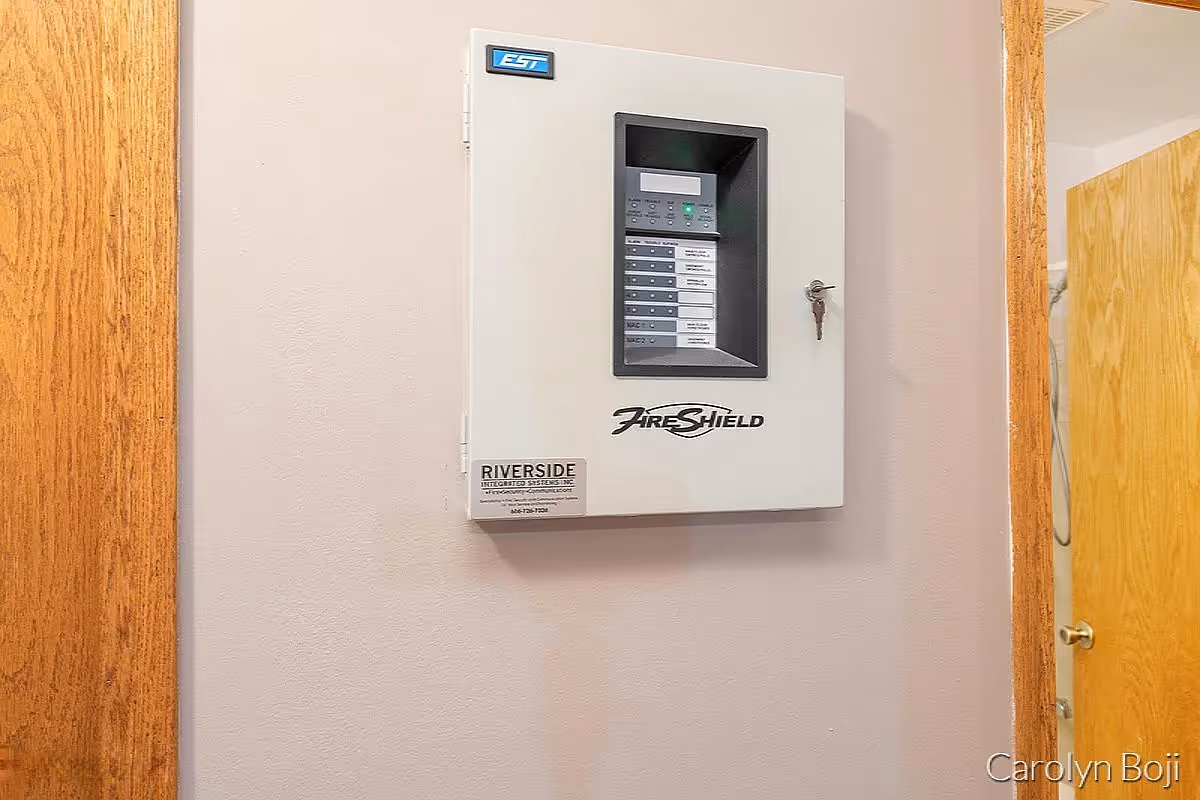 A wall-mounted fire alarm control panel labeled 'FireShield' with a key inserted in the lock, mounted on a beige wall between two wooden door frames.