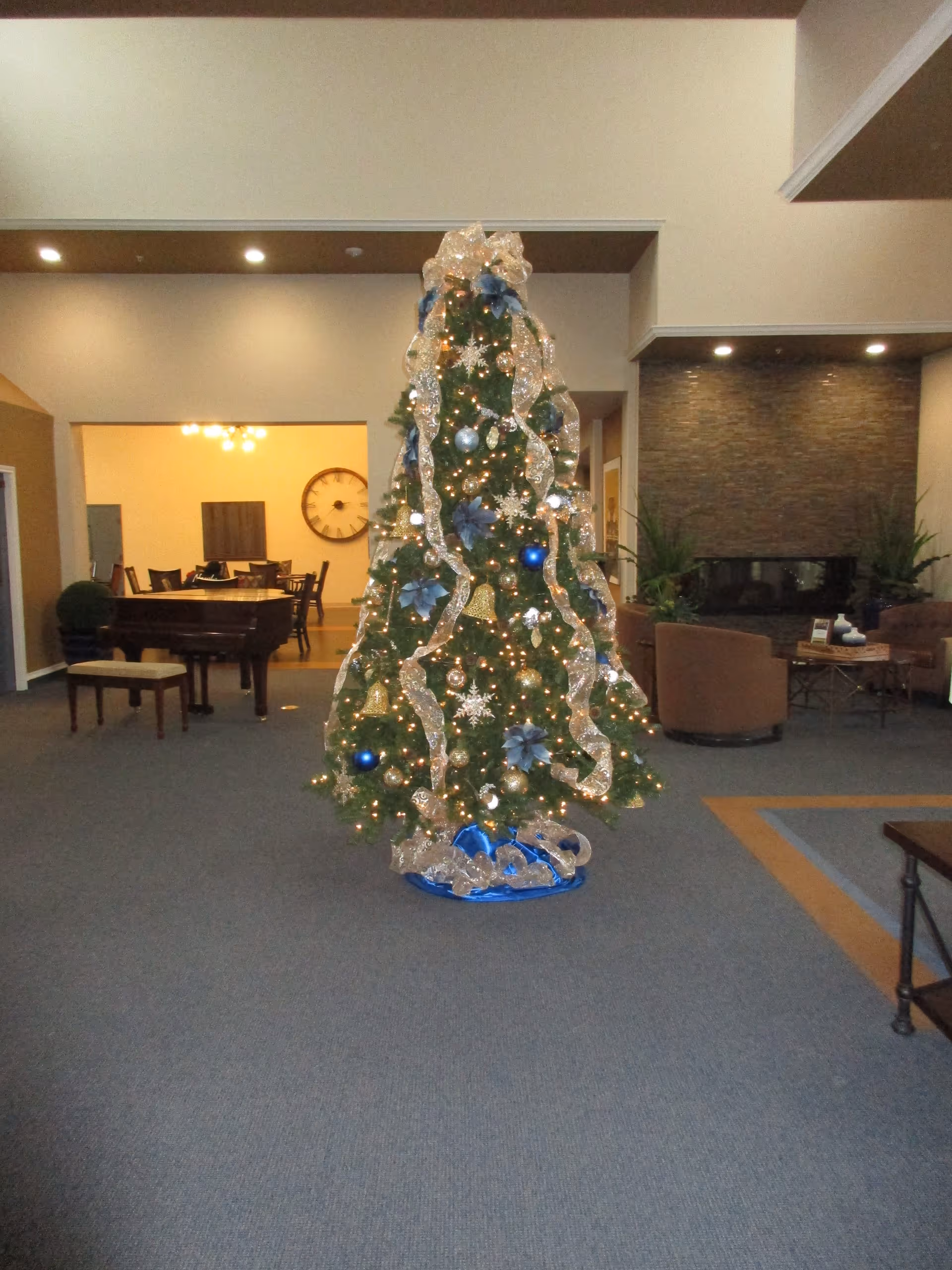 A decorated Christmas tree with gold ribbons, blue ornaments, and lights stands in the center of a spacious room. In the background, there is a grand piano with a bench, a large wall clock, a dining table with chairs, and a seating area with armchairs and a fireplace.