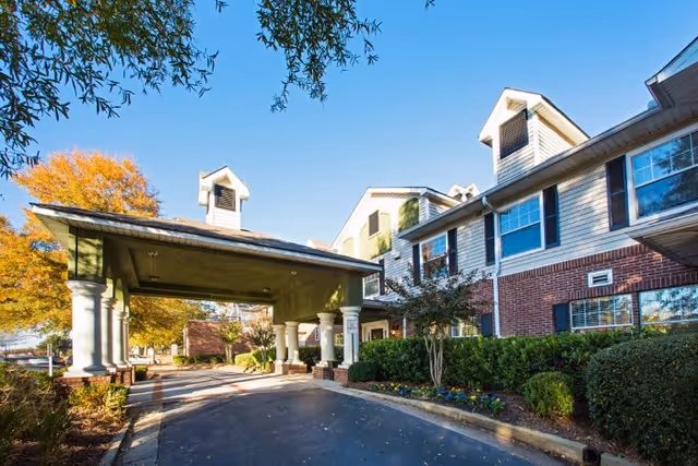 Exterior view of a senior living facility named Regal Pointe East Cobb, showing a covered driveway entrance with columns, well-maintained landscaping including bushes and flowers, and a clear blue sky.