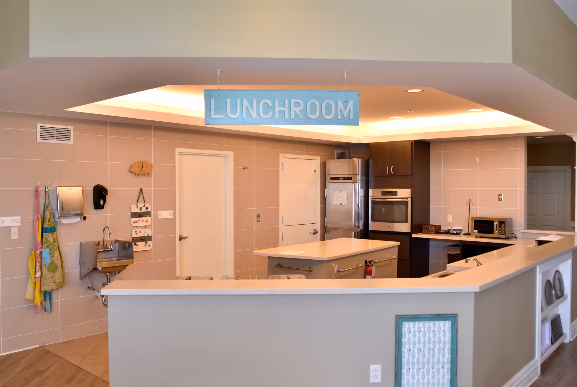 Interior view of a lunchroom in an assisted living facility featuring a kitchen area with a central island, stainless steel refrigerator and oven, microwave, and a sink. Aprons hang on the wall near a small handwashing sink. A blue sign hanging from the ceiling reads 'LUNCHROOM'.