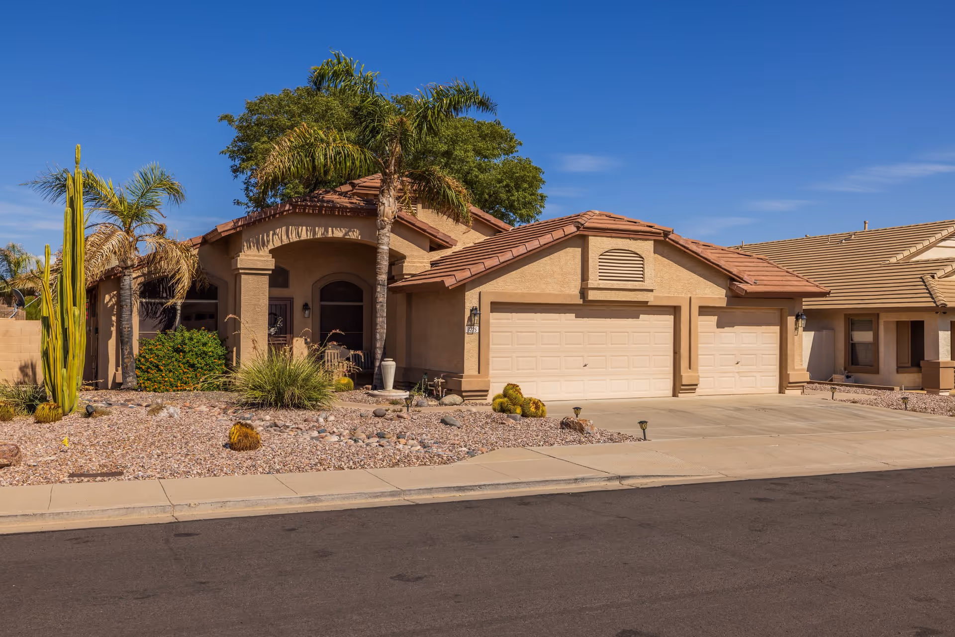 Exterior view of a single-story residential building with a three-car garage, desert landscaping with cacti and palm trees, and a clear blue sky.