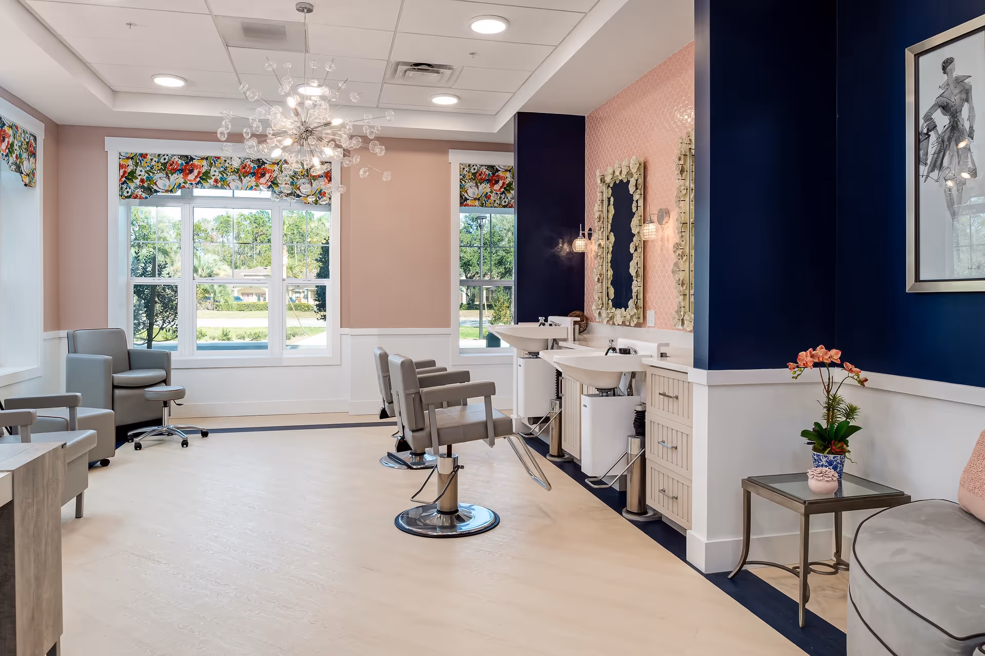 A bright and modern salon area with two styling chairs in front of sinks and mirrors, a comfortable seating area with armchairs and a small table with a flower pot, large windows with floral valances letting in natural light, and a decorative chandelier hanging from the ceiling.