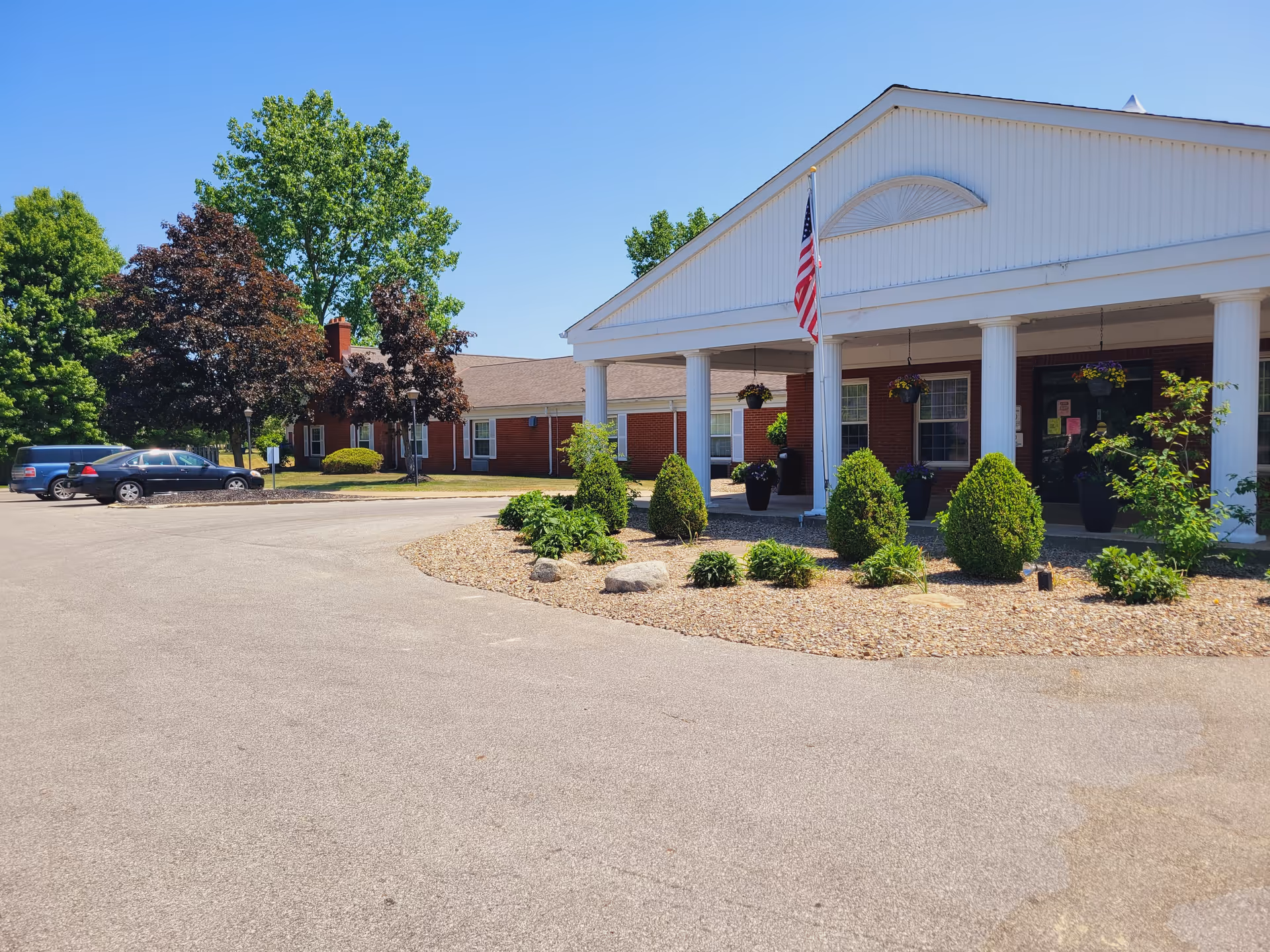 Exterior view of a single-story brick building with white columns and a covered entrance. There is an American flag hanging near the entrance, several potted plants, and neatly trimmed bushes in a landscaped area with rocks. Several cars are parked on the left side, and trees with green and reddish leaves are visible in the background under a clear blue sky.