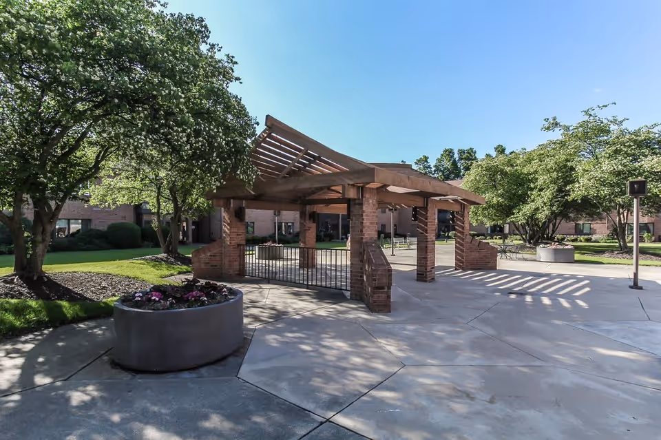 Outdoor courtyard area with a wooden pergola structure supported by brick pillars, surrounded by trees and large planters with flowers. The background shows a brick building under a clear blue sky.