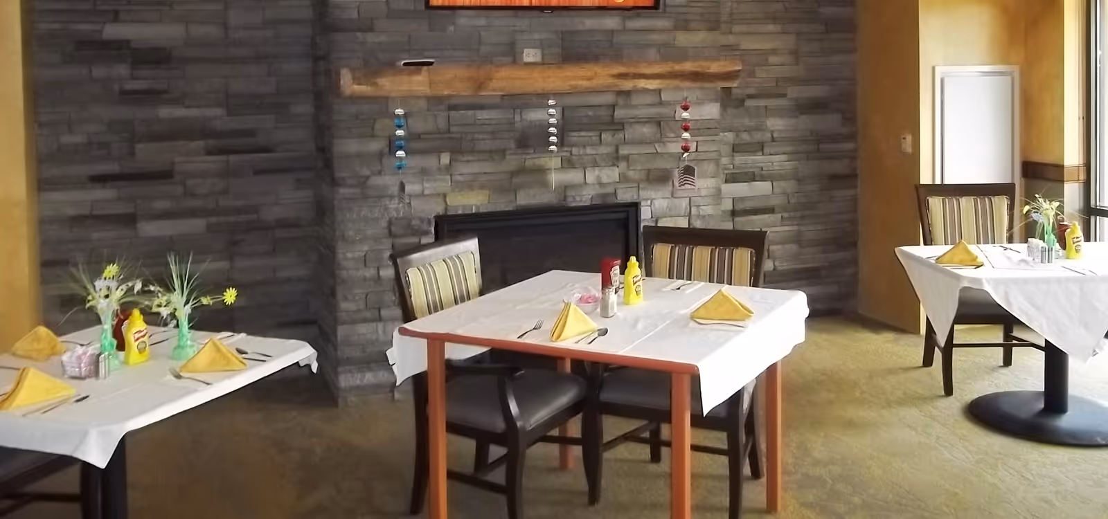 Dining room with several set tables and chairs, yellow napkins and condiments, in front of a stone fireplace wall.