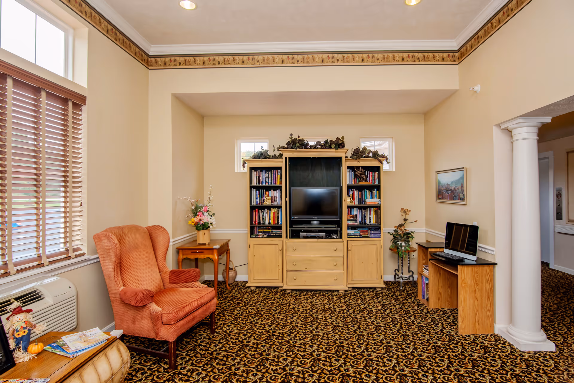 A cozy common room with an upholstered armchair, an entertainment center with a TV and bookshelves, a small computer desk, and patterned carpet.
