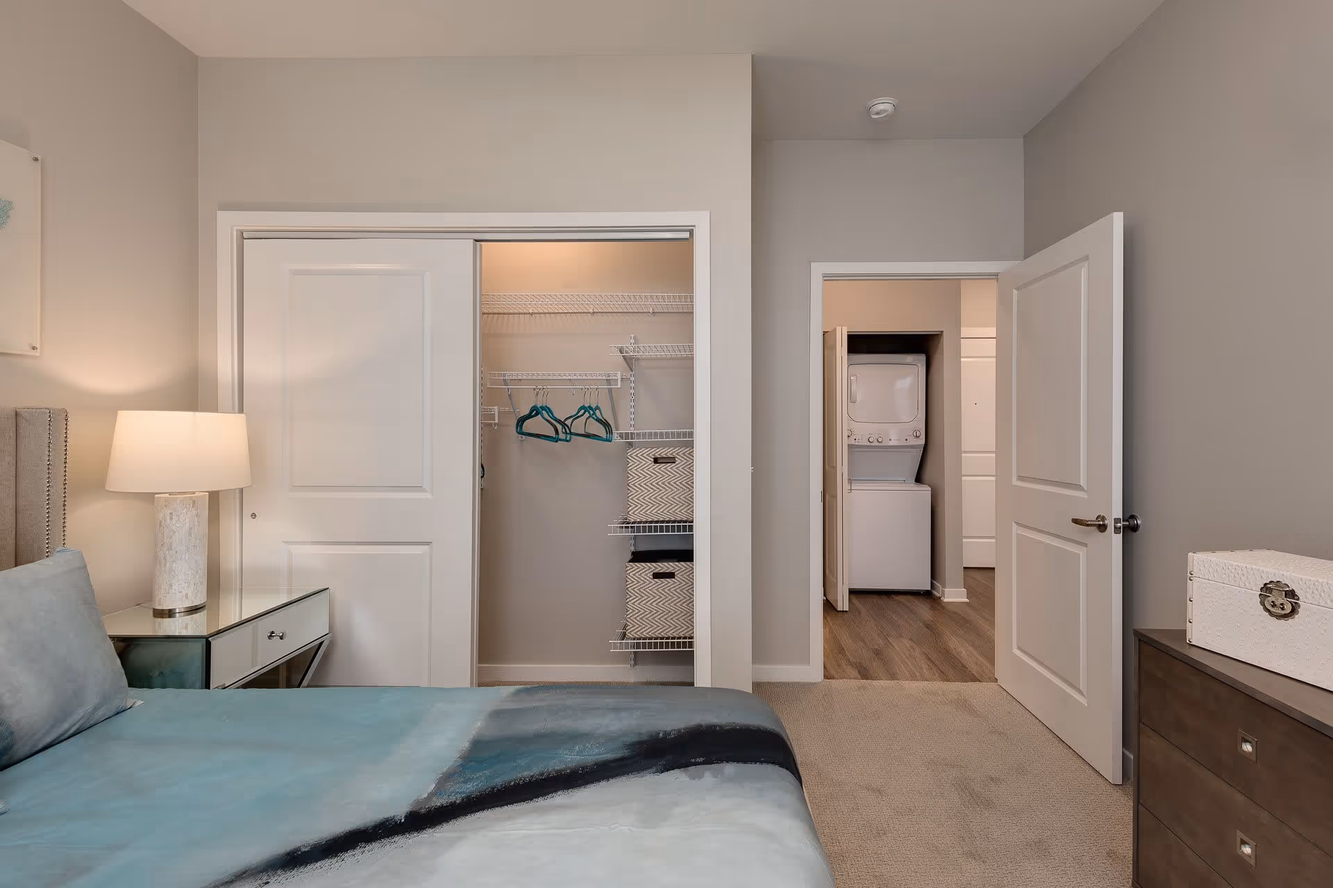 Bedroom with a made bed, nightstand and lamp, an open closet with shelving and hangers, and a doorway showing a stacked washer/dryer.