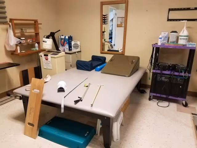A therapy or rehabilitation room with a large padded treatment table in the center, equipped with various physical therapy tools and supports. There is a wooden balance board leaning against the table, a step platform underneath, and a purple cart with medical supplies and equipment on the right. A mirror is mounted on the wall, and shelves with additional items are visible on the left side of the room.