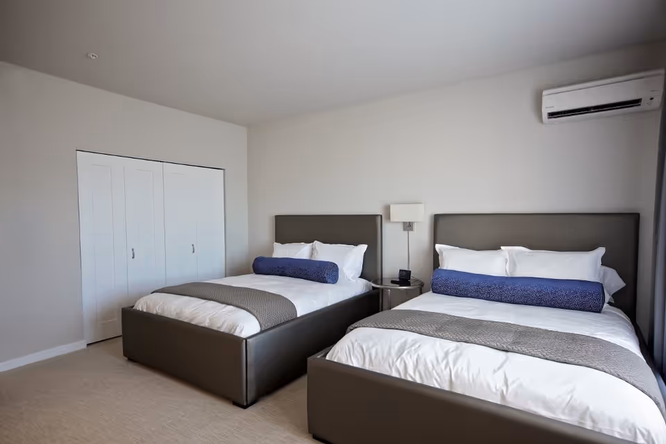 A bedroom with two beds featuring gray upholstered headboards and footboards. Each bed is made with white bedding, gray blankets at the foot, and long cylindrical blue pillows. Between the beds is a small round side table with a lamp mounted on the wall above it. There is a white closet with double doors on the left wall and a wall-mounted air conditioning unit on the right wall.