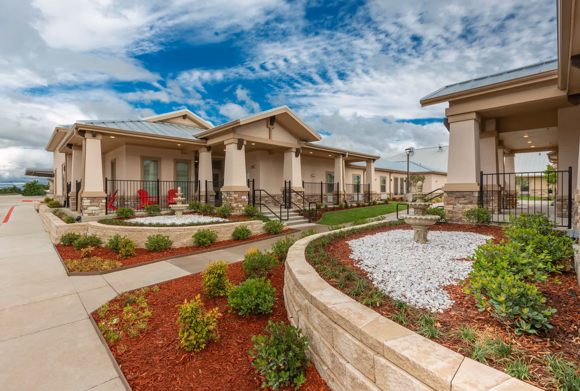 Exterior view of BeeHive Homes of Frisco showing a landscaped garden area with shrubs, mulch, and white decorative rocks surrounding a stone fountain. The building has beige walls, stone accents, and a metal roof with covered walkways and red chairs on a patio.