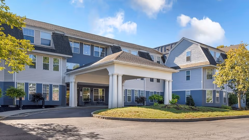 Exterior view of a multi-story senior living facility building with a covered entrance, surrounded by trees and landscaping under a partly cloudy sky.