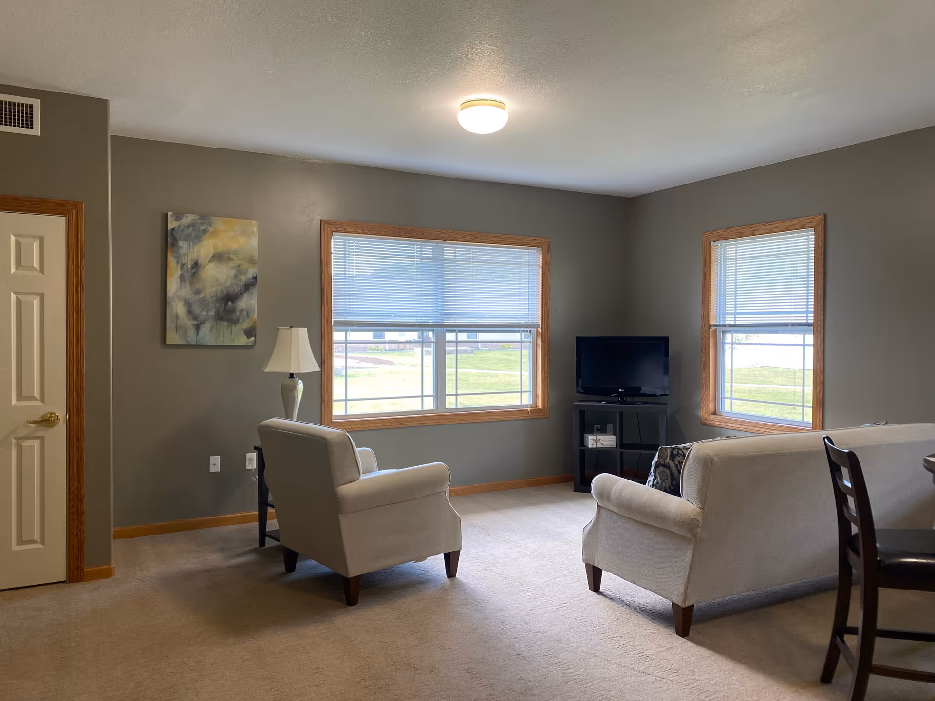 A living room with gray walls and beige carpet featuring a beige armchair and sofa, a small black TV stand with a flat-screen TV, two windows with blinds, a floor lamp, and a piece of abstract wall art.