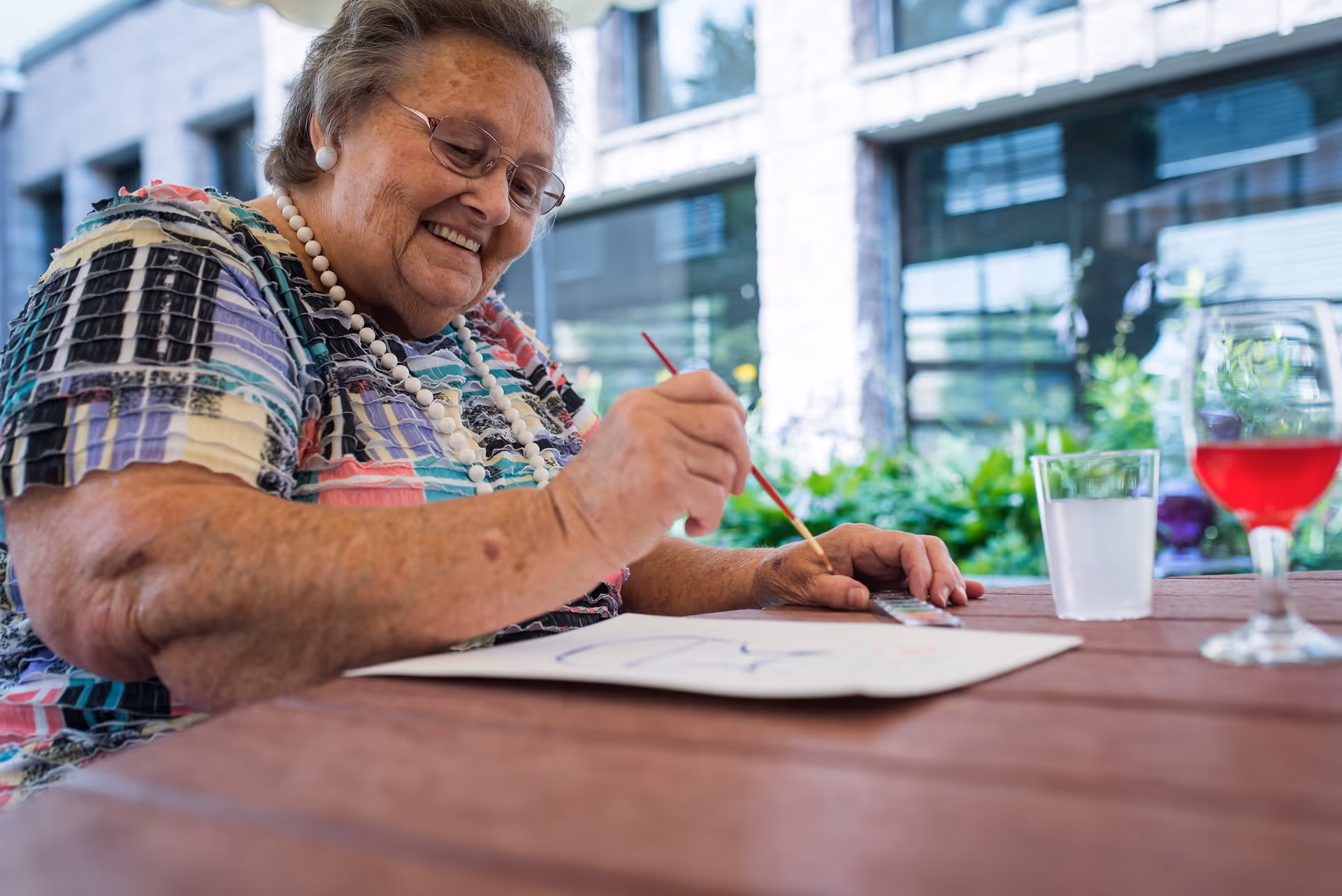 An elderly woman wearing glasses and a colorful patterned shirt is sitting at a wooden table outdoors, smiling as she paints on a piece of paper. There are two glasses on the table, one with water and one with a red beverage. The background shows a building with large windows and greenery.