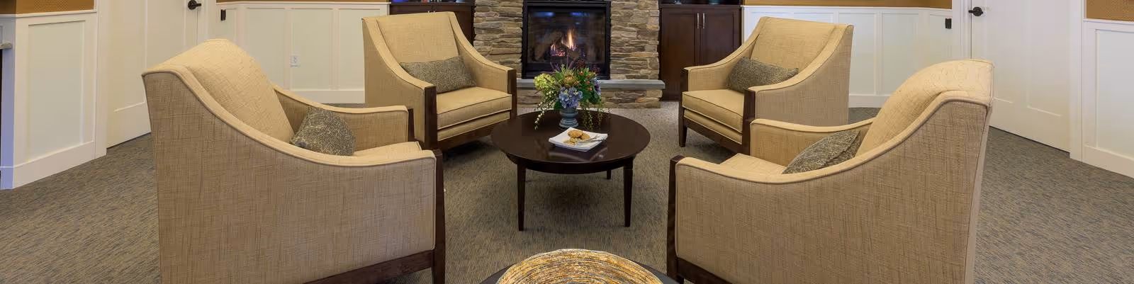 A cozy seating area with four beige armchairs arranged around a round dark wooden coffee table, which holds a small flower arrangement and a book. In the background, there is a stone fireplace with a fire burning, flanked by wooden cabinets. The room has light-colored walls with white wainscoting and carpeted flooring.