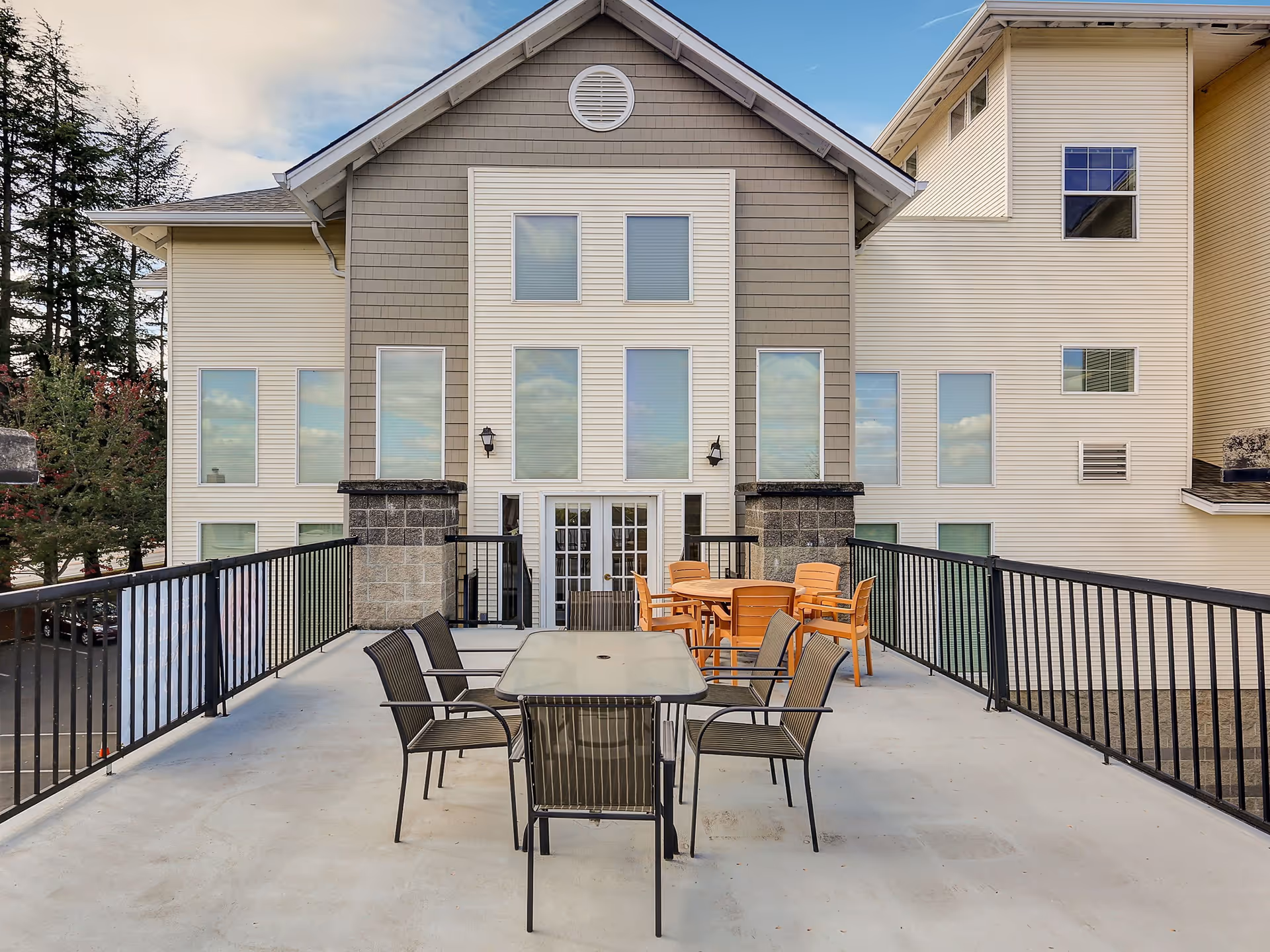 Outdoor patio area with two sets of tables and chairs on a concrete floor, surrounded by black metal railings. The patio is attached to a multi-story building with beige and gray siding and multiple windows. Trees and a partly cloudy sky are visible in the background.
