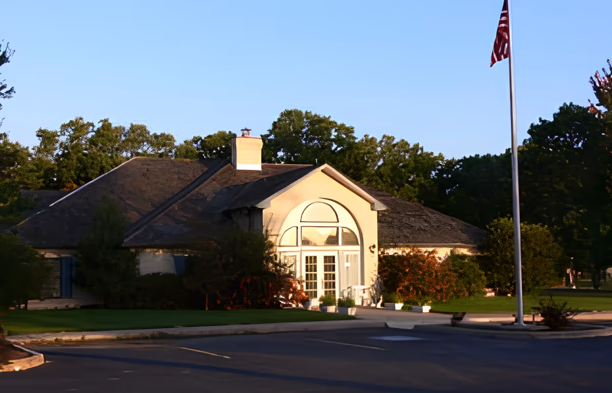 Front exterior of a single-story senior living building with an arched entryway and a flagpole on a landscaped lawn.