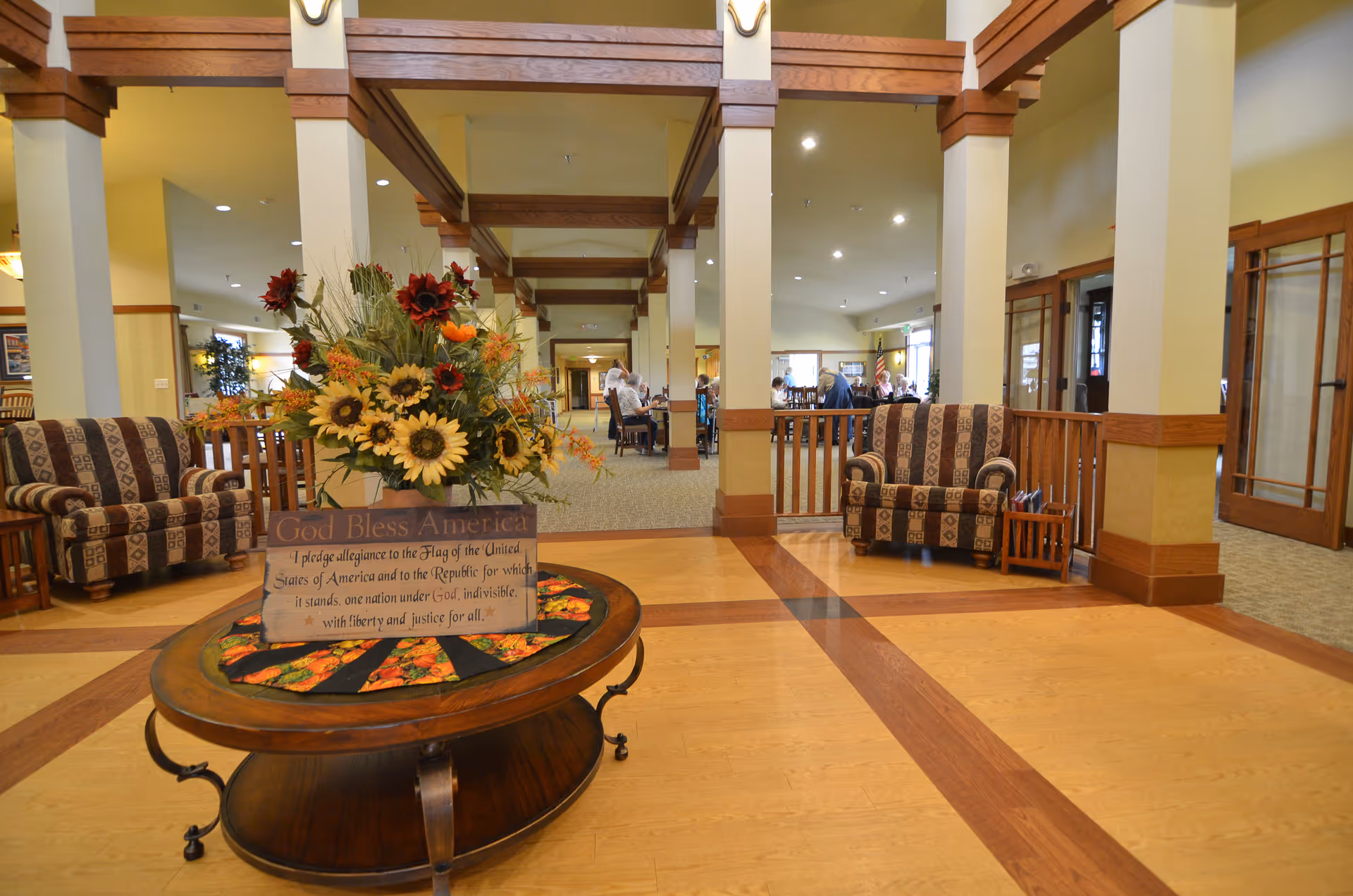 Spacious and well-lit common area in a senior living facility with wooden beams and columns. The room features patterned armchairs on either side, a round wooden table in the center with a floral arrangement and a sign that reads 'God Bless America' with a pledge text. In the background, several people are seated at tables in a dining or activity area.