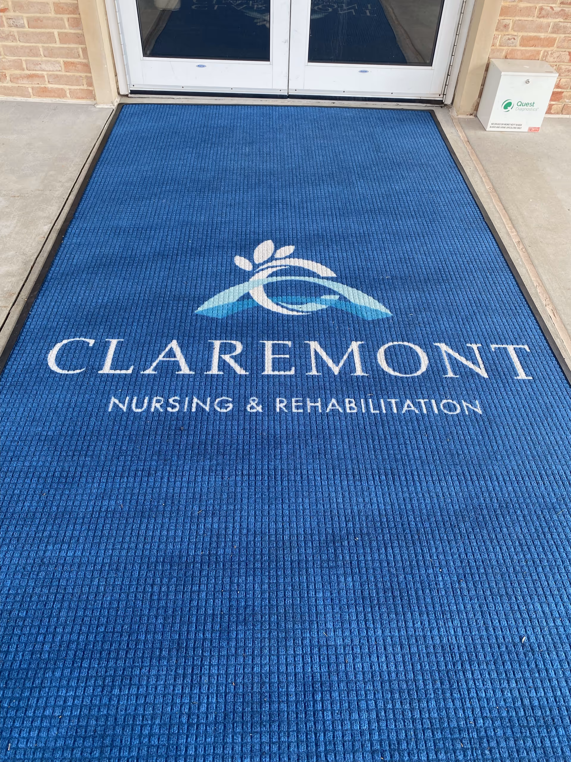 Blue entrance mat with the logo and text 'CLAREMONT NURSING & REHABILITATION' placed in front of glass double doors at the entrance of a building.