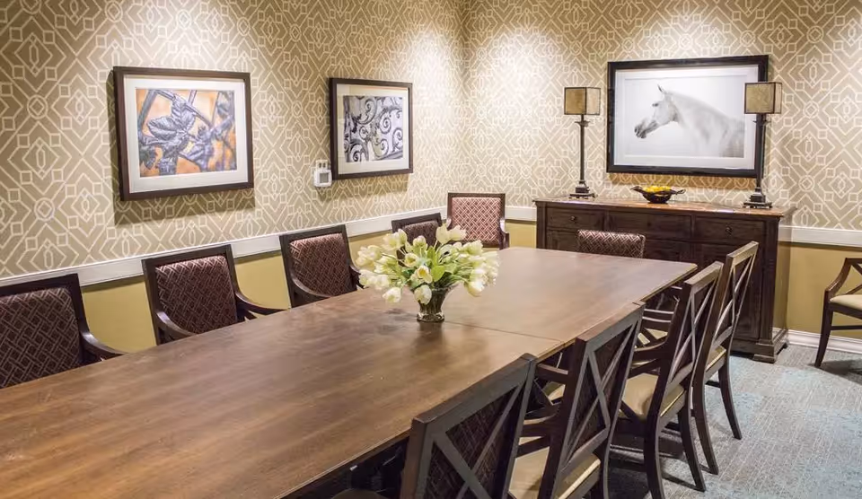A dining room with a long wooden table surrounded by chairs with patterned upholstery. A vase with white flowers is placed in the center of the table. The walls have a geometric patterned wallpaper and are decorated with framed artwork. A wooden sideboard with two lamps and a decorative bowl is against the far wall.
