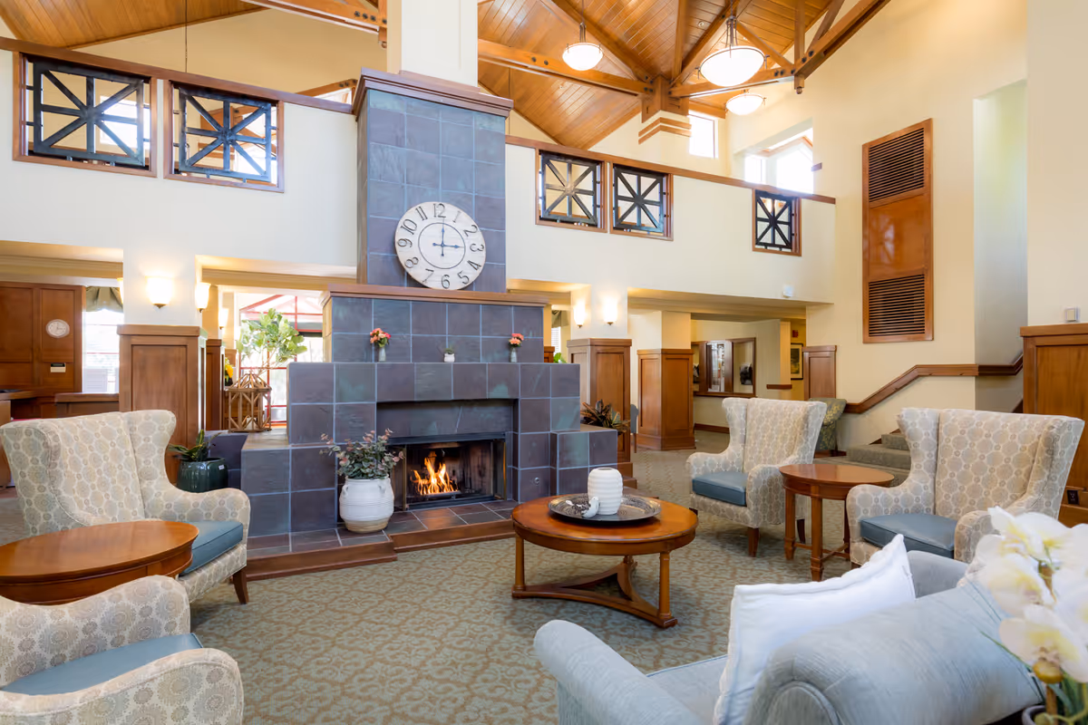 A spacious and well-lit living room area with a high wooden ceiling and a large tiled fireplace with a clock above it. The room features comfortable upholstered armchairs and sofas arranged around wooden coffee tables, with decorative plants and flowers placed on the tables and near the fireplace. The walls are painted in a warm beige tone, and there is a carpeted floor with a subtle pattern. The room has a cozy and inviting atmosphere.