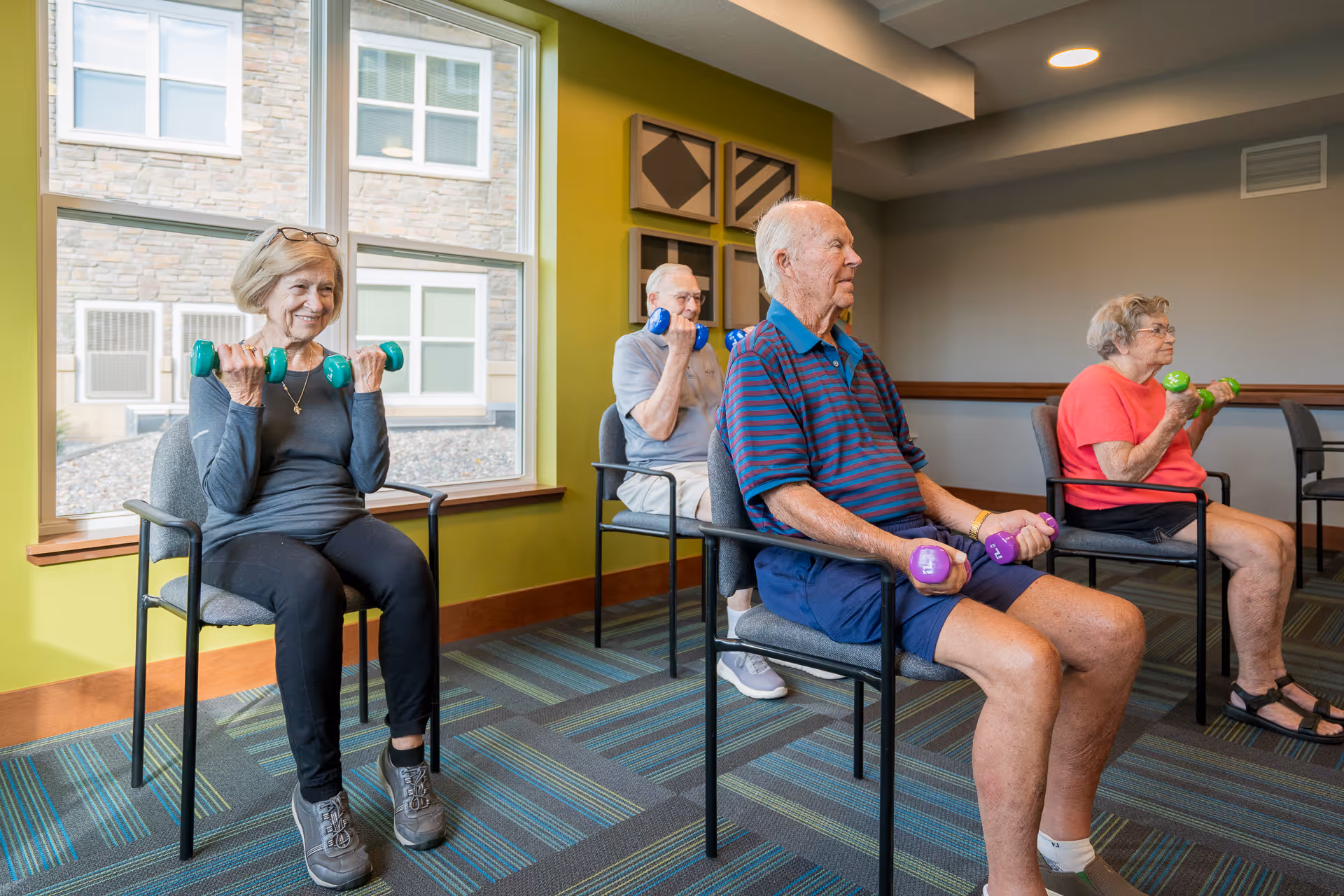 Four elderly individuals seated in chairs inside a room with large windows, participating in a light exercise session using colorful hand weights. The room has green and gray walls and patterned carpet flooring.