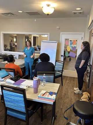 A group of people in a room at Iris Memory Care of NW Oklahoma City. One woman in medical scrubs is standing and speaking to two seated individuals. Another woman stands near a door decorated with colorful paper cutouts. A dog is sitting on the floor near a stool. The room has a table with books, papers, and a water bottle on it, and a large mirror on the wall.