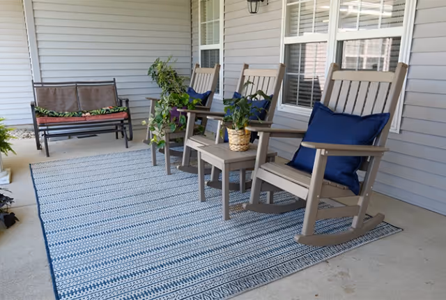 A covered outdoor patio area with a striped blue and white rug, two wooden rocking chairs with blue cushions, a small wooden table with a potted plant, and a bench with cushions against the wall of a building with gray siding and white-framed windows.