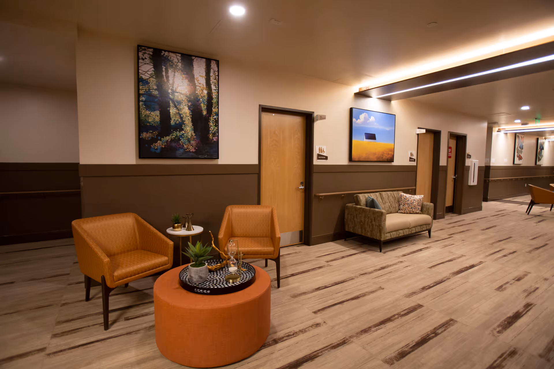 A well-lit hallway seating area in a senior living facility with two orange armchairs, a round orange ottoman with decorative items, a small white side table with plants, and a beige loveseat with pillows. The walls are decorated with colorful framed paintings, and there are wooden doors with restroom signs.